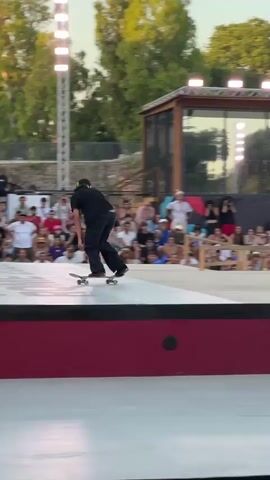 A male skateboarder in a black t-shirt, black pants, and a black baseball cap with headphones on is performing a trick on a skateboard ramp. The skateboarder is in mid-air, balancing on a rail that is part of the ramp structure. The background shows a crowd of spectators sitting on bleachers and standing behind a barrier, watching the event. Behind the spectators is a building with a wooden facade and large windows, illuminated by overhead lights. The environment appears to be an outdoor event with trees visible in the background. The event signage indicates "WORLD SKATE ROME." The video captures the skateboarder's action as he moves from one part of the ramp to another. The camera is positioned to capture the skateboarder in action from a side angle.