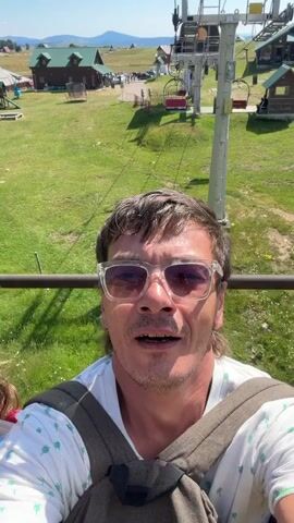 A man wearing a white t-shirt with a palm tree pattern and sunglasses takes a selfie while riding a ski lift. The ski lift is moving, indicated by the blurred background. He is smiling and appears to be enjoying the ride. The background shows a green grassy landscape with several wooden cabins and mountains in the distance under a clear blue sky.