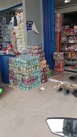 The video shows the exterior of a store. There are large stacks of goods, some of which appear to be packaged food items. There is also a blue barrel, a red barrel, a toy truck, and several pairs of sandals scattered on the ground. The store appears to be open, with blue accordion doors partially open. Shelving units filled with products are visible inside the store. A birdcage with a bird inside hangs from the ceiling. The overall scene suggests a retail environment with a significant amount of inventory displayed outside.