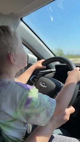 A young girl with blonde hair pulled back in a ponytail sits in the driver's seat of a car, holding the steering wheel with both hands. She is wearing a tie-dye shirt with purple, green, and white colors. An adult's hand is visible near the bottom of the frame, presumably assisting the girl. The car appears to be stationary on a dirt road on a sunny day. The dashboard is visible, showing digital information. Outside the window, there's a view of a field and a clear blue sky.