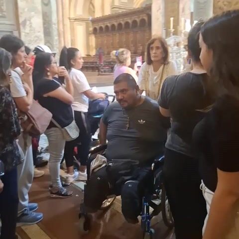 The video shows a group of people inside a large, ornate church or cathedral. A man in a wheelchair is in the foreground, surrounded by people who appear to be looking at something in the distance. The church has tall marble pillars and arches, and the altar is visible in the background. Several people are wearing casual clothing, and one person has a white hard hat on. The overall atmosphere seems calm and reverent.