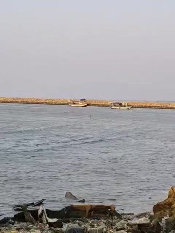 The video shows a body of water with several boats. In the foreground, there are some rocks and debris on the shore, including what appears to be a partially submerged, damaged wooden boat. Further out on the water, there are at least three fishing boats. These boats are moving and appear to be in the middle distance. In the background, there is a long, stone structure that looks like a breakwater or pier. The sky is overcast and a pale gray. The overall impression is a coastal scene, possibly a harbor or bay, with some signs of disrepair or neglect on the shore.