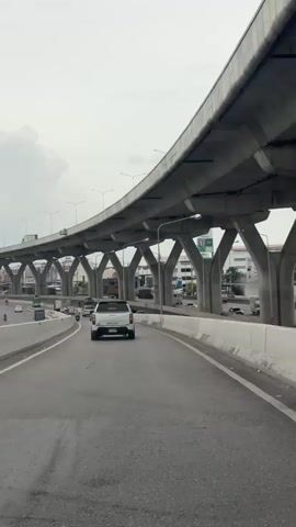 The video shows a view from a car driving on an elevated highway. The highway is made of concrete and supported by numerous concrete pillars. The sky is overcast. There are other vehicles on the highway, including cars, trucks, and a motorcycle. The road is asphalt. The car is traveling in the right lane. The video is a vertical shot.