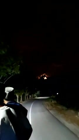 The video shows a person walking down a narrow, winding road at night. The road is paved with a white dividing line, and trees line either side. The sky is dark, but there is a faint reddish glow above the treeline. The person in the video is wearing a light-colored shirt and a hat. The footage is shaky, suggesting that it was filmed while in motion. Street lights are visible further down the road.