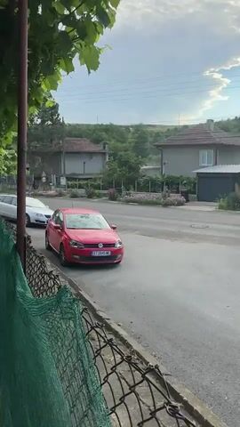 This media file captures a street view in Tsarevets, Bulgaria. The scene is set in a village, characterized by modest houses with red-tiled roofs and the presence of lush green trees and vegetation. The time of day appears to be late afternoon, as indicated by the lighting and the sky, which features a mix of white clouds and bright blue. The weather seems pleasant, with no signs of rain.

The primary focus of the video is the street and the vehicles on it. A red Volkswagen Polo is parked on the side of the road, its license plate visible as "BT 2845 ME". Further down the road, a silver station wagon is seen in motion, turning at an intersection. Another white vehicle is also visible in the distance. In the background, a few people can be seen walking, adding a sense of everyday life to the scene. One individual, wearing a red top, is walking near some houses.

Notable details include the electrical wires crisscrossing the sky, a common sight in rural areas. A street light is also visible. The foreground shows a grassy verge and a glimpse of a chain-link fence. The overall atmosphere is peaceful and tranquil, portraying a typical day in a Bulgarian village. The natural landscape, combined with the residential setting, creates a serene and somewhat rustic ambiance.