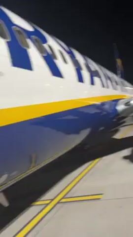 The video shows the side of a Ryanair airplane at night, followed by a view of someone ascending stairs to board the plane. The plane has a white top, a blue stripe along the middle, and the Ryanair logo in large blue letters. The stairs are gray with yellow accents. The setting appears to be an airport tarmac.