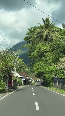 The video shows a drive down a paved road through a small village. The road has a white dividing line and is surrounded by houses and lush green trees, including palms.  Behind the village, a mountain rises, partially obscured by clouds. Several cars are seen driving on the road. The houses are simple structures with varying roof styles. Utility poles with wires run along the side of the road.