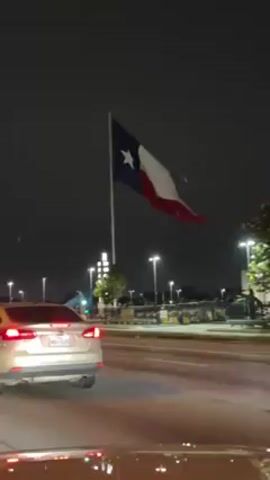 Here is a description of the video:

The video is taken from the interior of a car at night. The view is of a road, and a Texas flag waving in the wind. A gold car is in front of the filming car, waiting at a traffic light. Across the street is a John Deere dealership. There are street lights lining the road. The night sky is visible above. The overall lighting is dim and artificial due to the darkness and street lights.
