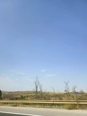 The video shows a view from a car, looking out at a dry, hilly landscape under a clear blue sky. A metal guardrail runs along the side of the road in the foreground. To the left, there are some trees, one of which appears bare and dead. In the distance, there are fields and what looks like a solar panel farm. The sun is shining brightly. The video is very short, lasting only three seconds.