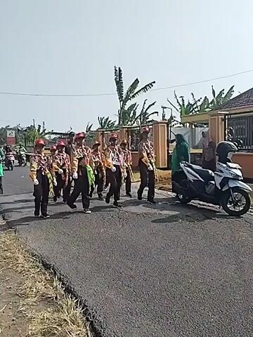 The video shows a group of young people, possibly scouts, marching in uniform on a paved road. They are wearing tan uniforms with brown hats, red accents, and colorful sashes. Some of them are also wearing face masks. They appear to be participating in a parade or a similar event. In the background, there are houses, trees, and a few motorcycles. The weather appears to be sunny and clear. The overall scene depicts an outdoor activity in a residential area.