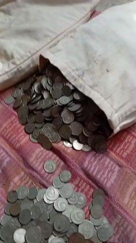 The video shows a pile of coins spilling out of a bag onto a patterned surface. The coins are small and silver-colored. The bag appears to be made of a light-colored fabric. Some of the coins have fallen onto the surface and are scattered around the bag. The scene is lit by natural light. The video is short and does not contain any audio.
