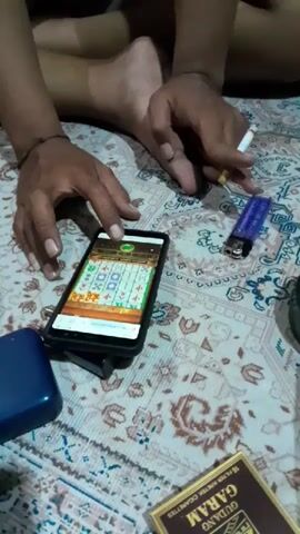 A person's hands are visible, one is holding a lit cigarette and a lighter, the other is touching a smartphone that displays what appears to be a gambling or lottery application. The person is seated on a patterned surface, possibly a bed or rug. In the foreground, a pack of cigarettes is visible. The lighting is dim.
