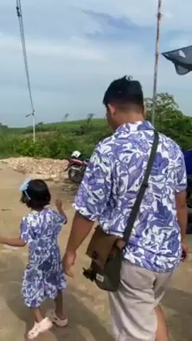 A man and a young girl, both wearing matching blue and white floral print outfits, walk down a dirt road towards parked cars and a cliff overlooking the ocean. The man has a brown messenger bag and is holding a phone. The girl has a blue bow in her hair. The sky is cloudy.