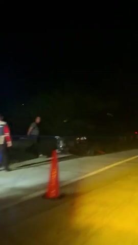 The video shows a nighttime scene on a road. A vehicle is visible, appearing to be stopped or moving slowly. The road has yellow and white lines. Lights from the vehicle and streetlights cast a yellow glow on the road. There are dark trees and foliage to the left of the road.