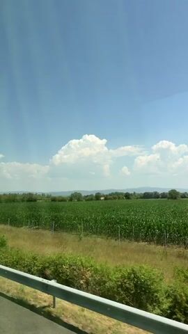 The video shows a view from a moving vehicle, looking out at a rural landscape under a bright blue sky with white clouds. In the foreground, there is a metal guardrail and a grassy area with some bushes. Beyond that, a large green field, possibly corn, stretches across the frame. Further in the distance, there are trees and hills. A blue and green irrigation system is visible in the middle ground. The scene appears to be a sunny day in the countryside.