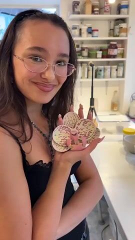 The video shows a young woman with long dark hair and glasses holding a pink and yellow ceramic orchid in her left hand. She is wearing a black tank top. In the background, there are shelves filled with jars and containers. To the right of the woman, there are several ceramic bowls and cups on a white counter. The camera then pans to a closer view of the ceramic pieces, showing a variety of bowls and cups, some of which are decorated with abstract designs. One bowl has a light blue glaze with a white decoration in the center, and another bowl is pink with a white, fish-shaped decoration. The video ends with a close-up of an empty pink bowl.