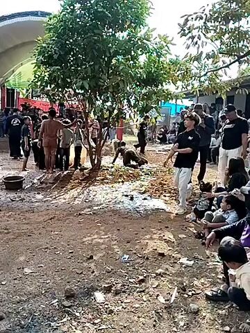 A group of people are gathered outdoors in what appears to be a village or rural setting. The ground is dirt and debris, with some puddles of water. There are trees and some simple structures in the background. In the foreground, a young person with curly hair is being filmed and appears to be dancing or posing. To the left, a group of people are standing, some watching the person in the foreground. To the right, other people are sitting or standing, also observing the scene. There is a general atmosphere of a community gathering or event.