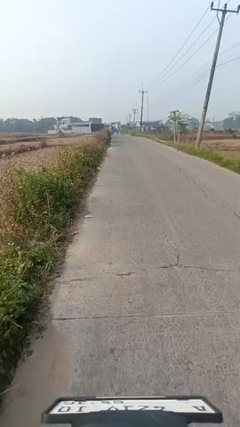 The video shows a first-person perspective of a person riding on a paved road in a rural area. The road is cracked and shows signs of wear. On the left side of the road, there are fields with dry grass and some vegetation. There are also some buildings visible in the distance. On the right side of the road, there are more fields, power lines, and utility poles. The sky is hazy and the overall lighting suggests it might be daytime. The video captures the movement of the rider, with occasional glimpses of a motorcycle's side mirror reflecting the surroundings. No other people or vehicles are clearly visible in the immediate vicinity.