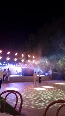 A video captures a nighttime outdoor event with a DJ setup on a stage. Multiple bright rectangular lights are positioned vertically above the DJ booth, emitting a pinkish-orange glow. Smoke or haze is present, illuminated by the stage lights. Two young boys are in the foreground, facing the stage and appearing to dance. One boy is wearing a light-colored outfit and the other a tan-colored outfit. A man in a white shirt is standing to the right of the stage, facing away from the camera, possibly taking a photo. Trees and foliage are visible in the dark background. The ground in the foreground is illuminated by scattered light patterns. The video appears to be a short recording of a moment at an outdoor party or concert.