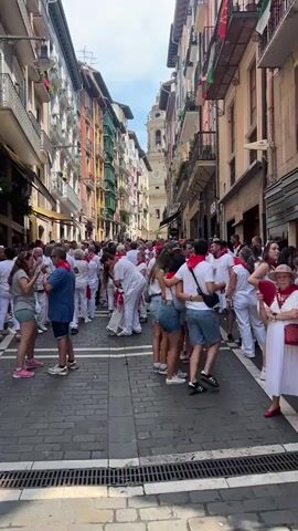 A crowded European street scene unfolds, likely during a festival given the attire. A large group of people, predominantly wearing white clothing and red neckerchiefs, fills a cobblestone street lined with multi-story buildings. The architecture suggests an old European city, with colorful facades and balconies. In the background, a tall building with a clock tower and a bell tower is visible. Some individuals are embracing, while others stand in groups, conversing or observing. The atmosphere appears festive and energetic. The street is paved with cobblestones and has a gutter with metal grates. The buildings have shops on the ground floor, some with awnings and advertising. The sky is partly cloudy. A digital thermometer mounted on a building shows a temperature of 39°C.