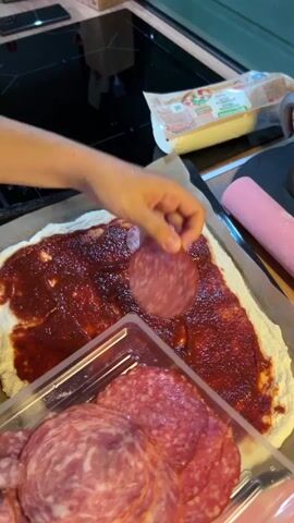 The video shows a close-up of a person's hands preparing a pizza. The pizza base is spread with a red sauce, likely tomato sauce, and a person is placing slices of salami on top of the sauce. In the foreground, a plastic container holds more pre-sliced salami. In the background, a package of cheese is visible, along with a pink rolling pin. The scene appears to be a kitchen setting.