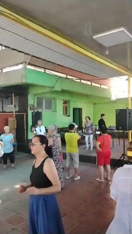 The video shows a group of people dancing outdoors. A woman in the foreground is wearing sunglasses and a blue skirt. She is looking to her left and appears to be dancing. In the background, several other people are dancing, some of whom are older. There is a stage set up with a woman singing into a microphone. A speaker is visible to the right of the stage. The environment appears to be a covered outdoor patio with green walls and a tiled floor. The lighting suggests it is daytime.