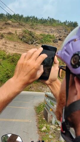 Here is a description of the video:
The video shows a person recording a hillside from a moving vehicle. The person is wearing a purple helmet and glasses and has a red beard. They are looking at their phone as they record a video. The hillside is covered in dirt and small patches of green vegetation. A thin stream of smoke rises from the top of the hill. A gray road stretches across the foreground, bordered by a metal guardrail. The weather appears overcast. The vehicle is moving along the road.