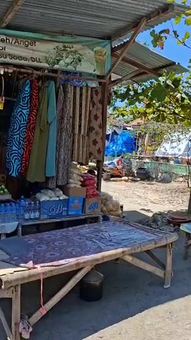 A street-level view pans across an outdoor market on a bright, sunny day. Stalls display various goods, including fabrics, bags of what appear to be food items, and what might be local crafts or souvenirs. A person sits at one of the stalls, appearing to be an attendant. The market is situated in what seems to be a coastal or tropical environment, with lush green trees and a clear blue sky. The overall atmosphere is relaxed and typical of a small, local marketplace.