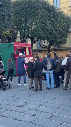 The video shows a small crowd gathered in an outdoor square. In the background is a small, green puppet stage with red curtains, where a puppet show appears to be taking place. People of various ages are standing and walking around, many facing the stage.  Some are in groups talking to each other.  A stroller is visible on the left.  The square is paved with gray stone. Tall trees are on the periphery, and yellow buildings with balconies form the backdrop.  The weather appears to be sunny.