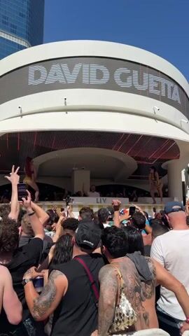 The video shows a DJ performance by David Guetta at the LIV Beach in Las Vegas. He is wearing a white t-shirt and has his arm raised, engaging with the crowd. Behind the DJ booth, two women are dancing. A large crowd is gathered, many holding up their phones to record the performance. The crowd consists of both men and women, and a few are shirtless. Tattoos are visible on several people. Some in the crowd are holding drinks. Overhead, the sign reads "DAVID GUETTA" in large, illuminated letters. The day is sunny and the sky is clear. A tall building is visible in the background.