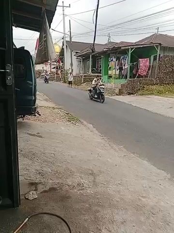 A person rides a scooter down a paved road. To the left, a parked van is visible. To the right, several houses with clothes drying outside are visible. Power lines stretch across the sky.