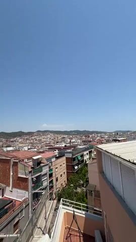 The video shows a panoramic view of a city from a high vantage point, likely a rooftop or balcony. The sky is clear and blue, indicating a sunny day. Below, a dense urban landscape unfolds, characterized by numerous buildings with terracotta roofs and brick facades. In the foreground, scaffolding with red and metal components is visible, suggesting construction or renovation work is in progress. The buildings vary in height and architectural style, forming a complex tapestry of urban development. In the distance, hills or mountains form a natural backdrop to the cityscape. The camera appears to be slowly panning or tilting, offering a sweeping perspective of the city. There are no people or animals clearly visible in the frame. The overall impression is that of a bustling city under a bright, clear sky.