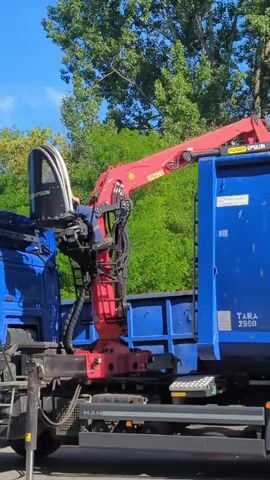 In a sunny outdoor scene, a large red hydraulic crane arm is extended over a blue skip on the back of a truck. The crane has an operator's cab, and a person is visible inside the cab. The crane arm itself has "EPSILON" and "GRIGG" text visible on it. The blue skip has "TARA 2980" and "CMG 101724" printed on it. The truck has "MAN" visible on the chassis. The background features lush green trees and a clear blue sky, suggesting a daytime setting. The overall impression is one of industrial activity, likely related to waste management or construction.