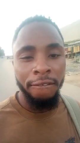 The video depicts a man with a beard and short, dark, curly hair walking outside, holding a phone up to record a selfie video. He's wearing a brown t-shirt with a graphic design and has a strap from a bag over his shoulder. The background transitions from what appears to be a sandy, dune-like environment to a more urban scene with buildings visible. At one point, the man makes an expression of surprise or shock, opening his eyes wide and pursing his lips. The video quality is somewhat shaky, and the lighting is bright.