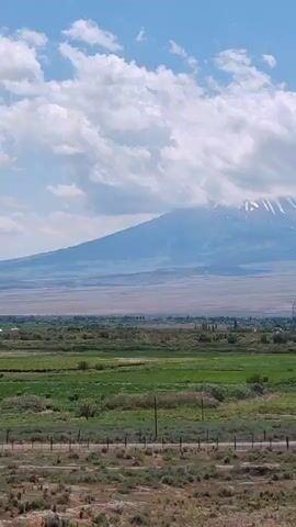 This is a vertical video of a scenic landscape. In the background, a large mountain range is visible, with some snow on the peaks. The sky is blue with white, fluffy clouds. In the middle ground, there is a vast, dry, dusty plain with a small town or village scattered across it. In the foreground, there is a field of green grass and other vegetation, with a fence and some utility poles. The camera pans slowly from left to right, revealing more of the landscape.