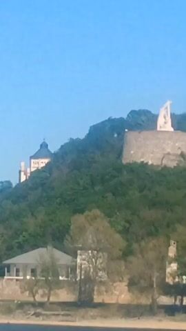 The video shows a view of a fort on a hill with a monument and a flag, followed by a view of a church with a dome. The fort is made of stone and surrounded by trees. The flag is the Hungarian flag. The church is made of stone and has a large dome. The sky is blue.