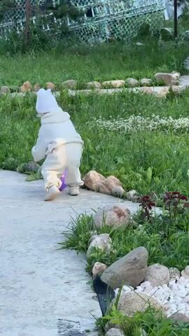 A toddler wearing a white hooded outfit and a white hat is playing with a purple ball on a concrete path outdoors. The toddler is initially crouched down, and then stands up and walks towards the left of the frame. The path is bordered by grass and rocks. In the background, there is more grass and some fencing.