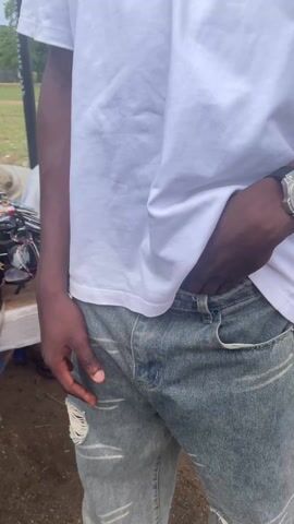 A person in a white t-shirt and ripped gray jeans is standing outdoors near a table of sunglasses. The person raises their right hand towards the camera, revealing the palm of their hand. The person’s left hand remains in the left front pocket of the jeans. A large, silver watch is on the person’s left wrist. The ground appears sandy.