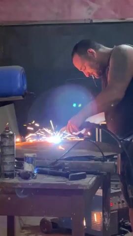 A man with a beard and tattoos is seen in a workshop, standing at a wooden workbench. He is engaged in welding, holding a welding torch in his right hand and a metal rod in his left, which he feeds into the weld point. Bright sparks and intense light emanate from the welding process on the workbench. A cigarette is visible in his mouth. Various tools and objects, including a gas canister, cans, and a hammer, are scattered on the workbench. A blue plastic container is positioned on a shelf above the workbench. The background is a dark wall, and the workshop appears to be dimly lit.