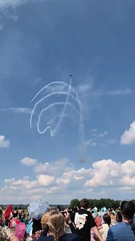 The video shows an outdoor event with spectators, possibly an airshow. In the sky, three airplanes are performing aerobatics, leaving white contrails that form a swirling pattern against a blue sky with scattered clouds. The spectators in the foreground are mostly seen from the back, with some heads and shoulders visible. They appear to be watching the aerial display. The overall environment suggests a clear, sunny day suitable for an outdoor event.