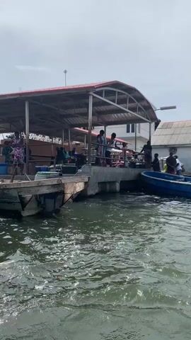 The video shows a waterfront scene with people and boats. There is a covered dock area with people standing and moving around on it. Several boats are docked nearby, including a blue one and a partially visible wooden boat. The water is choppy. In the distance, there are buildings. Later, a long, narrow boat filled with people passes by. The boat is being propelled by a person using a long pole. More buildings are visible in the background. The sky is overcast.