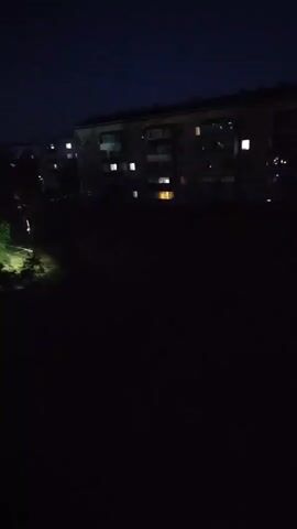 This media file is a short video clip taken at night from an elevated position, likely a window, overlooking a residential area. The scene is dark, with the exception of artificial lights. In the foreground, the edge of a wooden window frame is visible on the left.

The middle ground of the scene features a patch of green foliage, illuminated by a bright light source, possibly a streetlamp or car headlights. Beyond this, a line of parked cars is visible, their headlights adding to the scattered illumination. Further in the background, several multi-story buildings are silhouetted against the night sky. Lights are visible in the windows of these buildings, indicating occupancy. The sky is a deep blue, and a bright, full moon is prominent in the upper left quadrant of the frame, casting a soft glow.

The overall impression is a quiet, urban night scene. The time of day is clearly night due to the darkness and artificial lighting. The weather appears clear, as evidenced by the visible moon. There are no people or overt activities taking place, suggesting a moment of stillness. The location cues, such as the style of buildings and the presence of a Cyrillic script on the timestamp which indicates it was taken in Lisakovsk, Kazakhstan. The mood is tranquil and perhaps a little melancholic due to the darkness and stillness. There is no visible text in the media itself, other than the timestamp overlay.