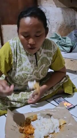 A woman with dark hair is sitting on the floor, eating a meal. She is wearing a green and white patterned dress. She is using a spoon to eat rice and what appears to be some kind of stir-fry or dish with sauce. In front of her on the floor is a piece of brown paper with a mound of white rice, some food with red sauce, and a small yellow item, possibly a piece of fruit. A smartphone is lying on the floor to her right, displaying an image or video. The background is somewhat dimly lit and appears to be an indoor space with a textured wall.