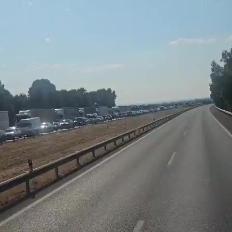 The video shows a long line of traffic backed up on a highway under a clear, hazy sky. The camera is positioned from inside a vehicle moving along the highway, providing a view of the stationary vehicles in the adjacent lane. The traffic jam consists of numerous cars and trucks, stretching far into the distance. The highway is paved and has a metal guardrail separating it from the shoulder and the stationary traffic. Trees and foliage are visible on the left side of the highway, and on the right side, the highway curves away, with a tall tree visible in the distance. The lighting suggests it is daytime, possibly late morning or afternoon, given the haziness in the air. There are no people clearly visible within the stationary vehicles due to the distance and the video's quality. The overall impression is one of significant traffic congestion.