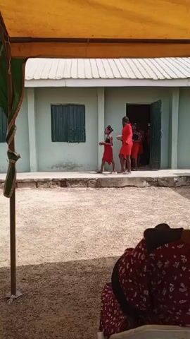 A group of young girls dressed in red dresses are walking in a line outside a building. A man in white stands in the doorway. The girls are walking on a concrete path in front of a light green building.  The scene is partially shaded by a yellow and green canopy, with a person seated in the foreground. The ground is light colored and appears to be gravel.