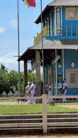 A group of people in white robes are walking towards a blue building with a multi-tiered roof. Some of the people are carrying musical instruments. The building has intricate white decorations and a small yellow flag on a pole. There is a fence in the foreground. The sky is blue with some clouds.