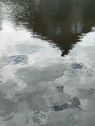 The video displays a reflection on the surface of water. The reflection shows a building with a dark roof and windows, as well as the reflection of clouds in the sky. The water is rippling, causing the reflections to appear distorted. The overall scene is monochromatic, with shades of gray and dark brown dominating. No people or specific actions are visible. The environment appears to be outdoors, with the water likely being a lake, river, or pond.