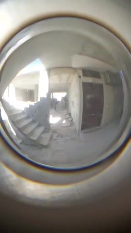 The interior of an unfinished building is viewed through a fisheye lens. There are concrete stairs and a closed door visible. Debris is scattered on the floor. Natural light enters from an opening at the far end of the room. A person is visible in the distance, but obscured by the bright light.