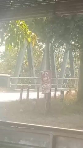 The video shows a close-up of a sign attached to a wooden post. The sign is red and white and appears to be advertising something. In the background, there is a bridge with large concrete supports. Trees and foliage are visible around the bridge and the sign. The lighting suggests it is daytime, possibly morning or late afternoon, with the sun casting a bright glare. The camera is positioned behind a wooden structure, possibly a window or railing, which frames the bottom of the shot. No people are visible in the frame.