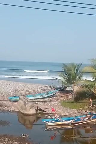 The video shows a bright, sunny day at a beach. The ocean has moderate waves with whitecaps. Several small, colorful fishing boats are pulled up onto the shore. Some of the boats are blue and white, while others are blue and yellow. They are propped up on wooden stands. A few palm trees are visible, their fronds swaying gently in the breeze. The ground is a mix of sand, pebbles, and some debris. There are power lines strung across the top of the frame, obscuring a portion of the sky.